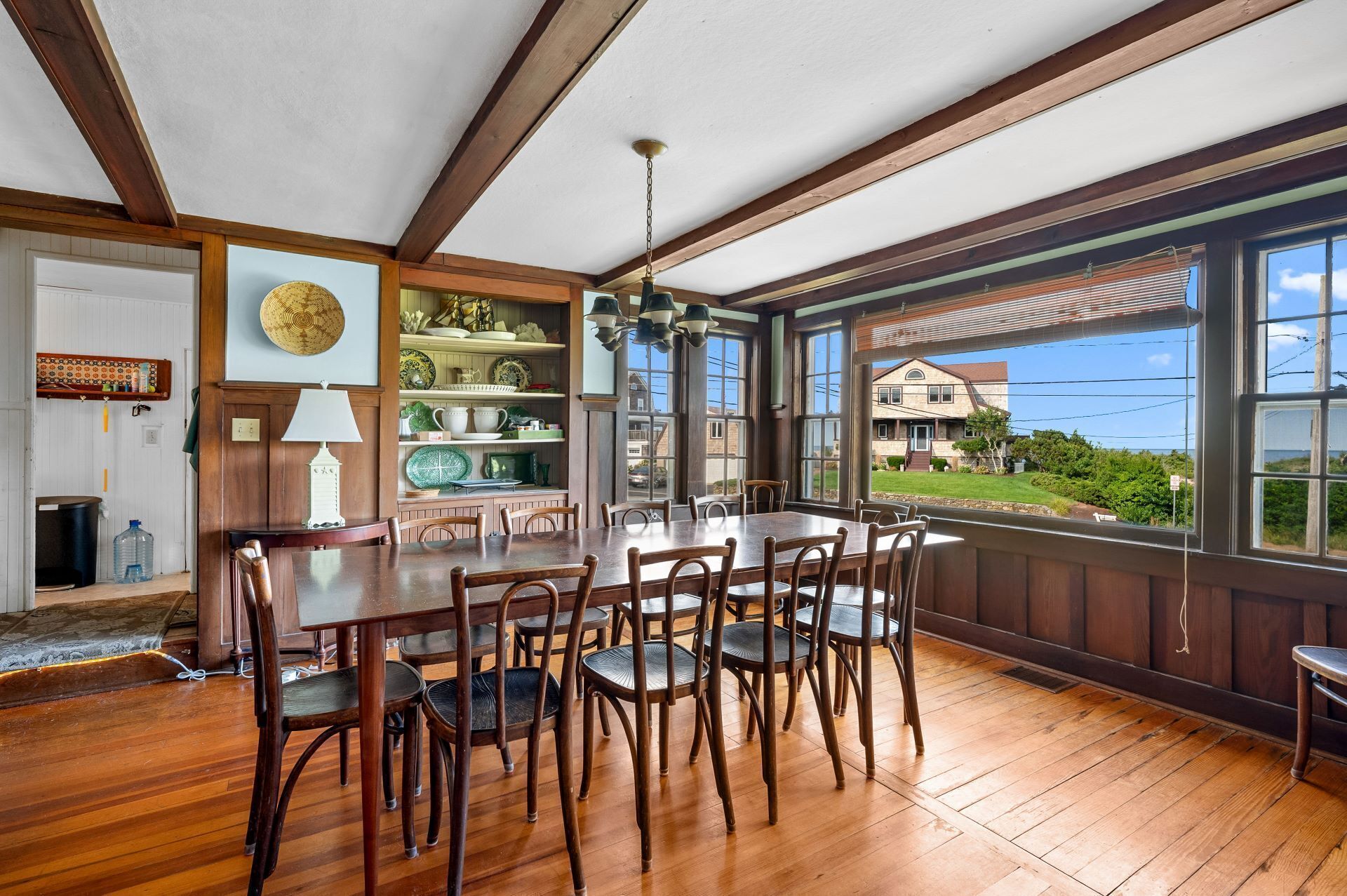 A dining area with a wooden table and chairs, a hanging light fixture, and a view of a house through large windows.