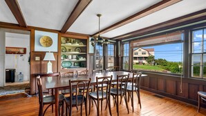 Un comedor con una mesa y sillas de madera, una lámpara colgante y vistas a una casa a través de amplios ventanales.