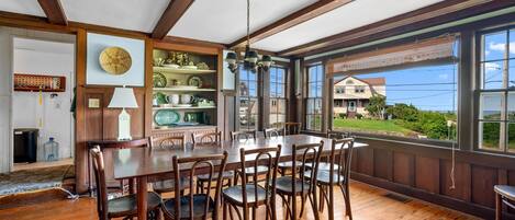 A dining area with a wooden table and chairs, a hanging light fixture, and a view of a house through large windows.