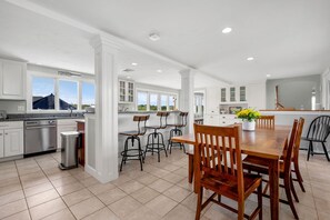 A spacious kitchen with white cabinetry, a dining area with wooden chairs, and a view of the outdoors through large windows.