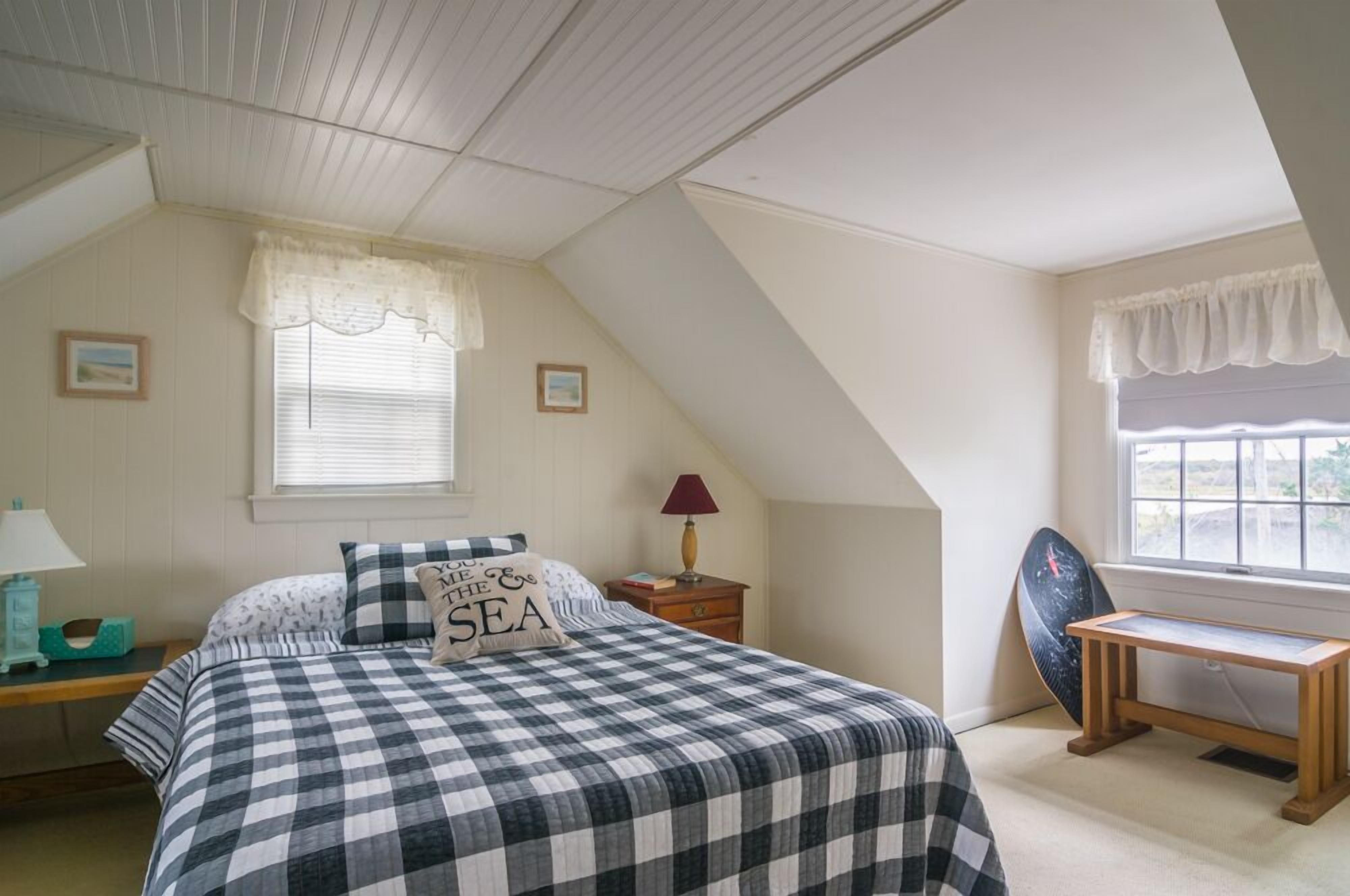 Une chambre avec une couvre-lit à carreaux, une table de chevet avec une lampe, une fenêtre donnant sur un beau paysage et une planche de surf appuyée contre le mur.