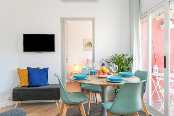 Uma sala de estar moderna com televisão de tela plana, um sofá com almofadas, uma mesa de jantar com cadeiras azuis e uma varanda com guarda-corpo.