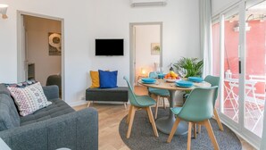 A modern living room with a grey sofa, teal chairs, a dining table with blue plates, and a TV mounted on the wall.