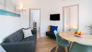 A modern living room with a sofa, a dining table with chairs, a flat-screen TV, and a painting on the wall.