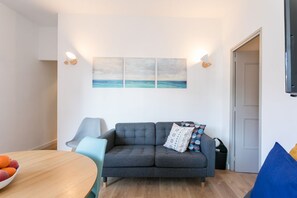Uma sala de estar moderna com um sofá cinzento, uma mesa de jantar de madeira com frutas e uma obra de arte na parede retratando uma cena de praia.