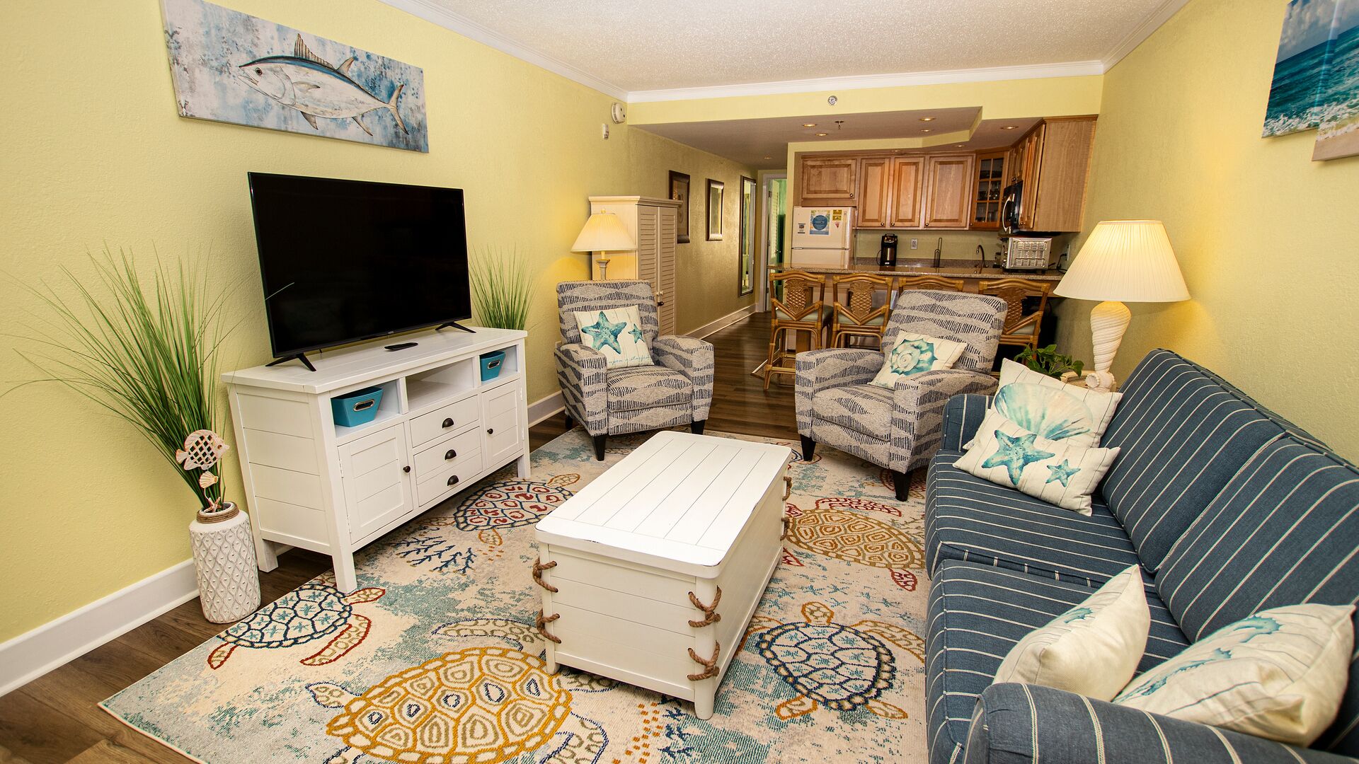 A living room with a television, two armchairs, a sofa, and a coffee table. There is a kitchen in the background.