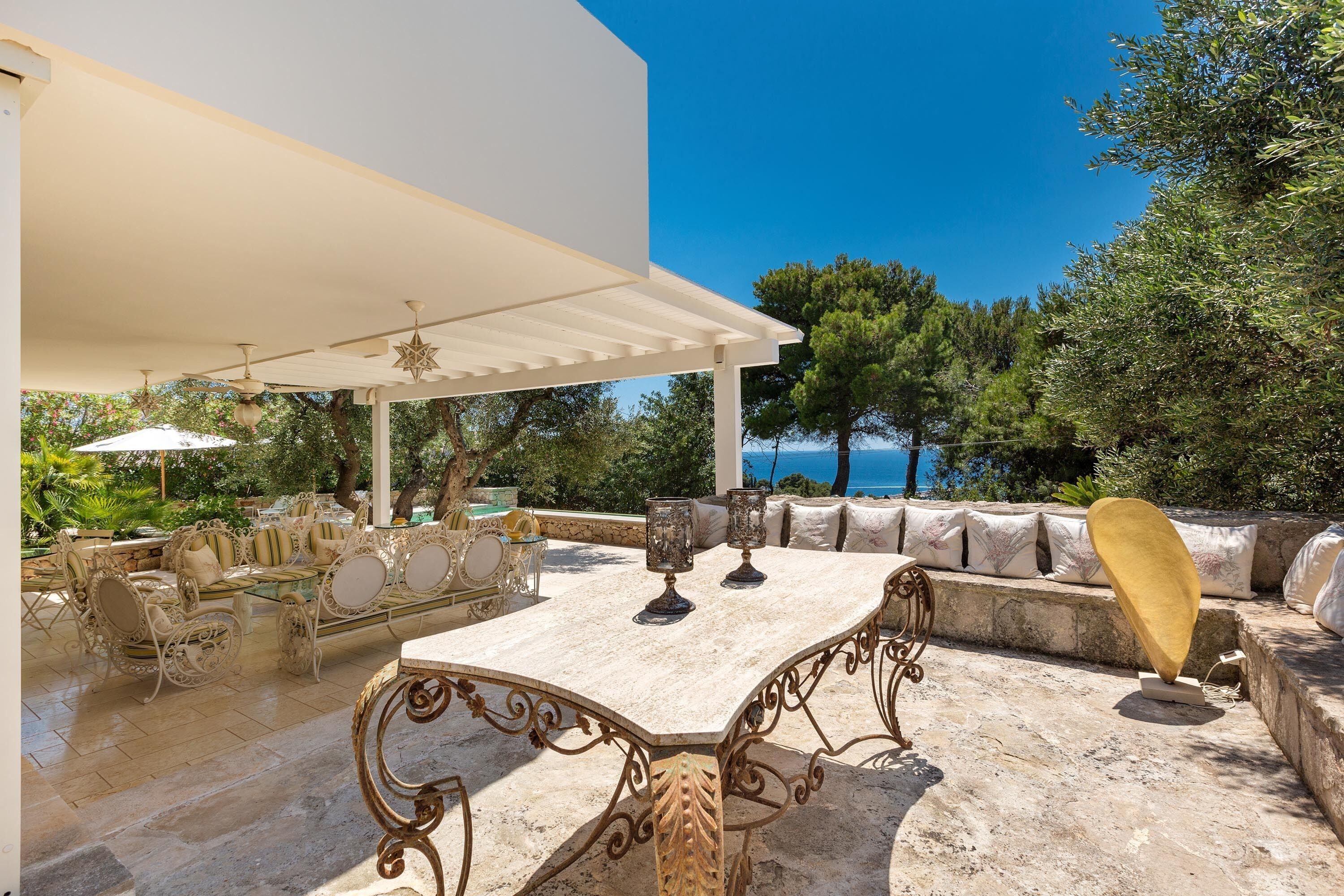 An outdoor patio with a white pergola, a marble table, and a seating area with cushions and a sofa.