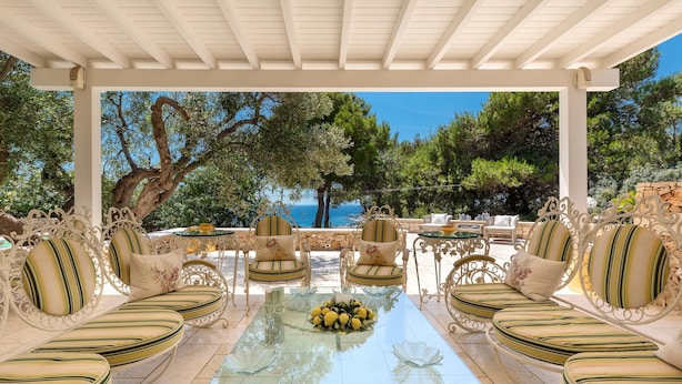 An outdoor patio with white furniture, a glass table with lemons, and a view of the sea and trees. - 2457 Villa Allegra by Perle di Puglia (Castrignano del Capo)