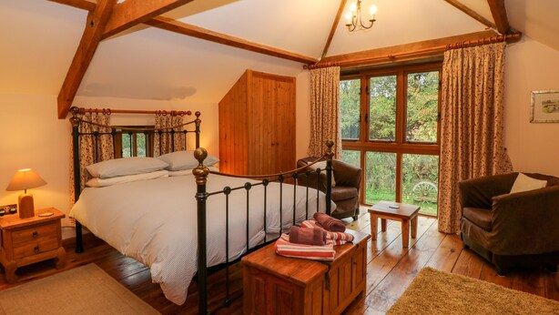 Un dormitorio con cama de madera, cómoda de madera, una silla de madera, una mesita pequeña, una mesita de noche y una ventana con cortinas.