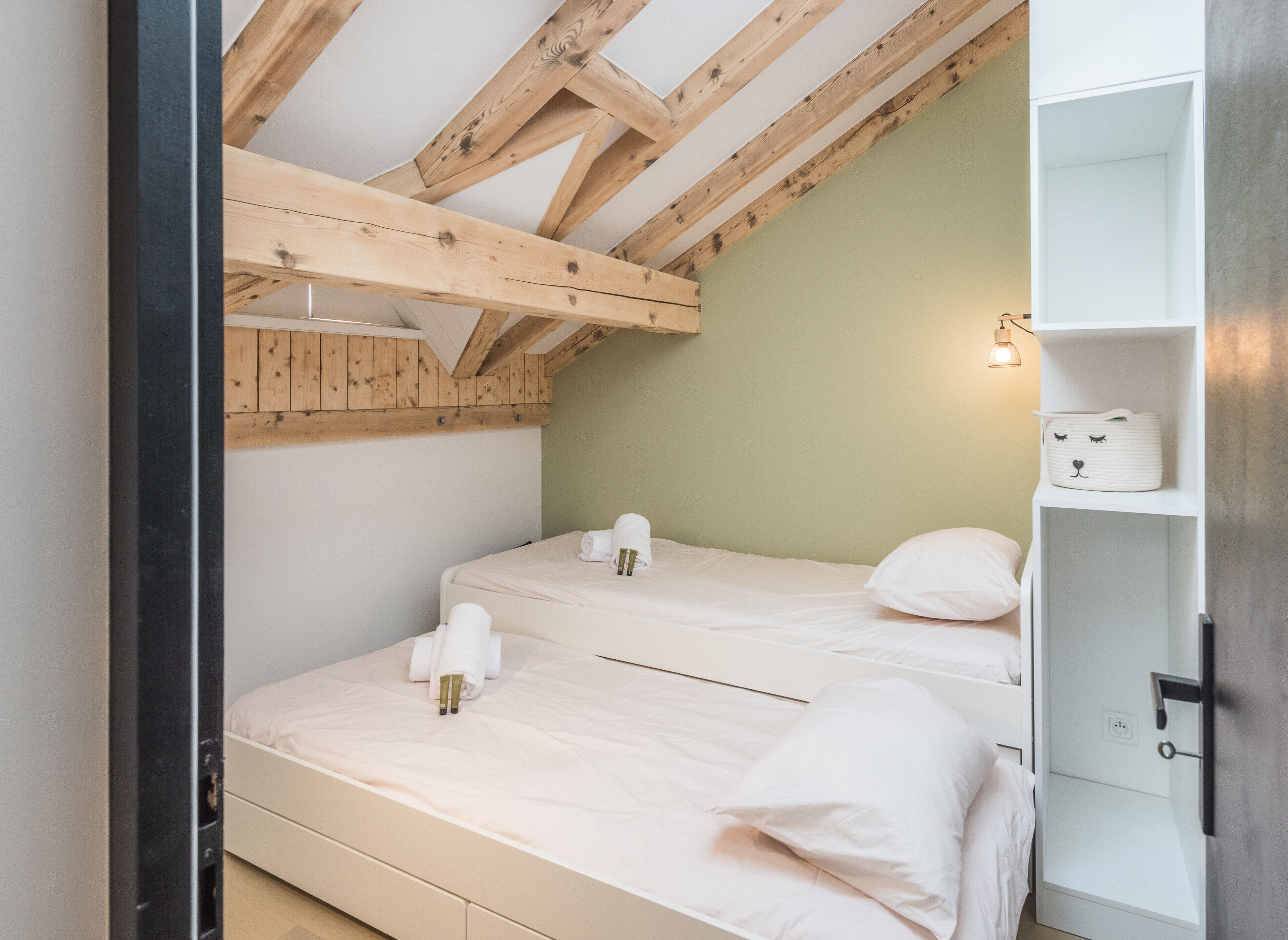 Une chambre avec deux lits, des poutres en bois au plafond, un mur vert et une armoire blanche ornée d’un motif représentant un visage de chat.