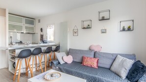Een moderne woonkamer met een grijze bank, zwarte stoelen, een witte salontafel, en een keuken met een raam.