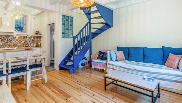 A spacious living area with a wooden floor, a blue staircase, a dining table with chairs, and a sofa with cushions.