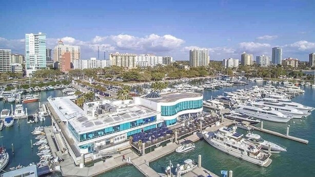 Un puerto deportivo con varias embarcaciones amarradas, un edificio moderno con techo curvo y un horizonte urbano al fondo.
