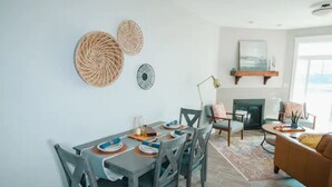 Un comedor con una mesa dispuesta para cuatro personas, rodeada de sillas. Se aprecia una chimenea al fondo y un sofá a la derecha.