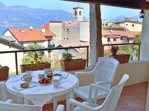 A balcony with a table set for two, potted plants, and a view of buildings and mountains.