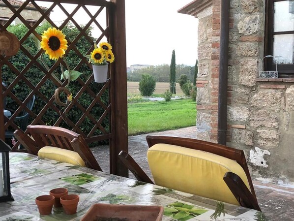 A patio with a table set for two, a wooden chair, and a yellow cushion. There are potted plants and a lattice structure with sunflowers.