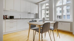Une cuisine moderne avec des meubles blancs, une table à manger entourée de quatre chaises et de grandes fenêtres donnant sur les bâtiments environnants.