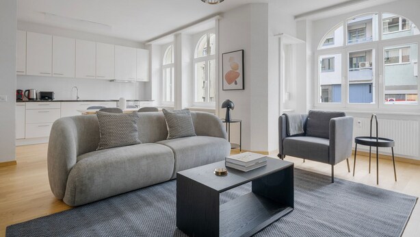 Un salon moderne avec un canapé gris, une table basse et un fauteuil. On aperçoit une cuisine en arrière-plan, avec des meubles blancs et une fenêtre.