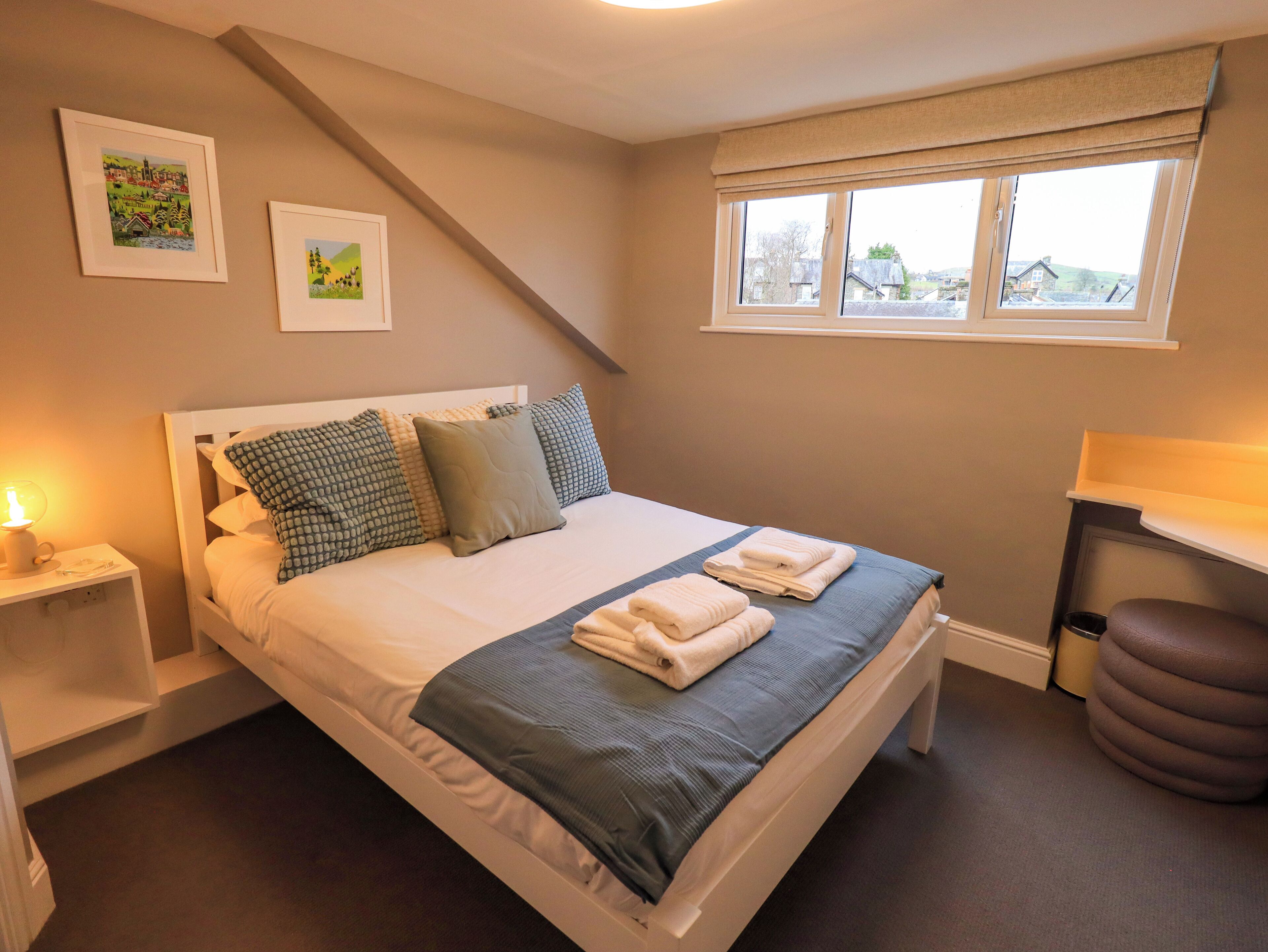 A neatly made bed with pillows, a bedside table with a lamp, and a window with a view of buildings.