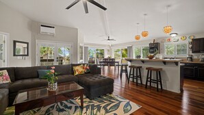 A spacious living room with a dark-colored sofa, a glass coffee table, and a large island with stools. There is a ceiling fan, pendant lights, and a view of the outdoors through large windows. - STEPS FROM STUNNING MOLOAA BAY (Anahola)
