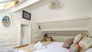 Un dormitorio con techo inclinado, cama con ropa blanca, varios almohadones, una silla colgante y una mesita de noche con lámpara.