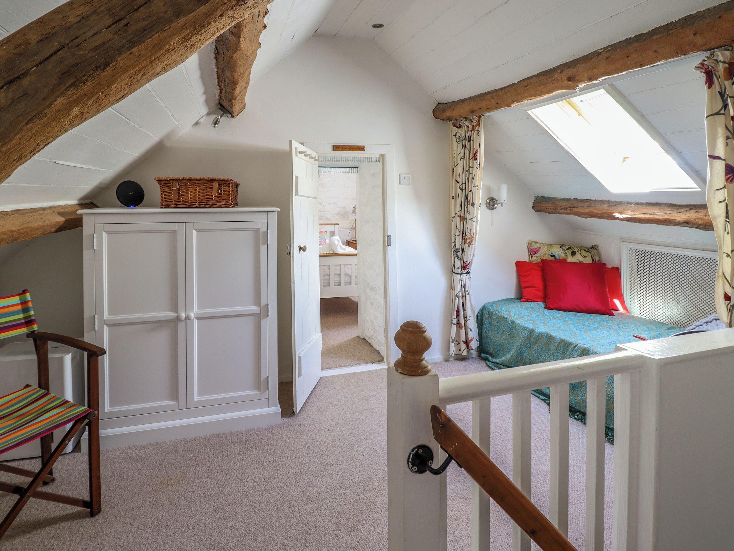 A bedroom with a sloped ceiling, a bed with red pillows, a white wardrobe, and a chair with a striped cushion.
