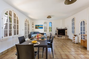 A dining area with a table set for four, surrounded by wicker chairs, a fireplace, and arched windows.