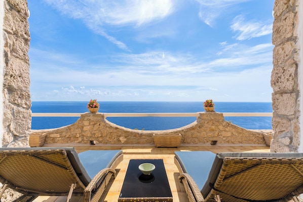 A terrace with stone walls, two wicker chairs, a table with a bowl, and a view of the sea.