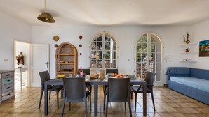 Ein Essbereich mit Tisch für vier Personen, umgeben von Korbstühlen, einem blauen Sofa und Blick durch Bogenfenster.
