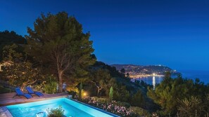 A pool area with lounge chairs, surrounded by trees and a view of a cityscape at night.