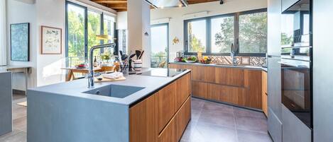 A modern kitchen with a central island, wooden cabinets, and large windows offering views of the outdoors.