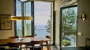 A dining area with a wooden table and chairs, large windows, and a view of the sea.
