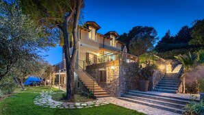 A two-story house with a stone staircase leading to a balcony, surrounded by greenery and trees.
