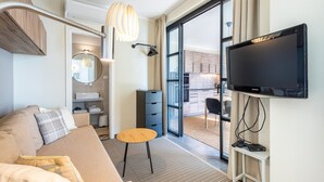A modern hotel room with a sofa, a small round table, a TV, a wardrobe, and a bathroom visible through an open door.