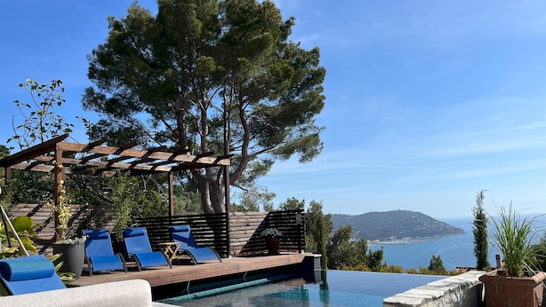 An infinity pool with a wooden deck and lounge chairs, surrounded by potted plants and a pergola.