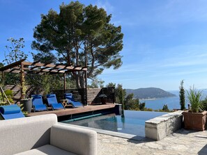 An infinity pool with a wooden deck and lounge chairs, surrounded by potted plants and a pergola. - Villa Nel Blu 8 (Cervo)
