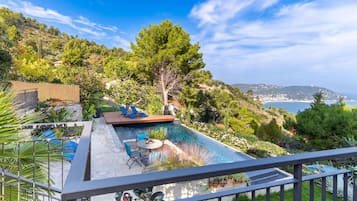 Una piscina infinita en la azotea con vistas al mar, rodeada de exuberante vegetación y una terraza de madera con sillas de descanso.
