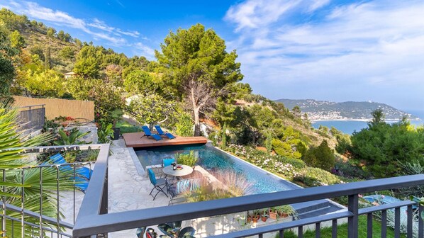 A rooftop infinity pool with a view of the sea, surrounded by lush greenery and a wooden deck with lounge chairs.