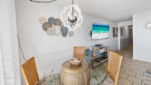 A dining area with a glass table, wicker chairs, a decorative bowl, a chandelier, wall art, and a flat-screen TV. - Eastern Shores - 101 1 Bedroom Condo by RedAwning (Santa Rosa Beach)