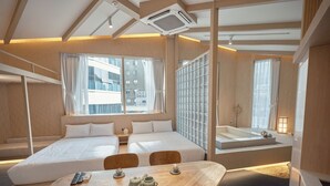 Habitación de hotel moderna con una cama grande, una mesa de comedor de madera con vajillas de té y vistas a la ciudad a través de amplios ventanales.