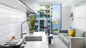 A modern kitchen with white tiles, a toaster, and a sink. There is a balcony with a view of a building and a body of water. A gray sofa with a yellow pillow is in the living area. - Modern High-Rise Condo with Pool/Gym, in Central DT MIAMI! (Miami)