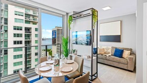 A modern living room with a large window, a dining area with a round table, and a sofa with cushions. - Modern High-Rise Condo with Pool/Gym, in Central DT MIAMI! (Miami)