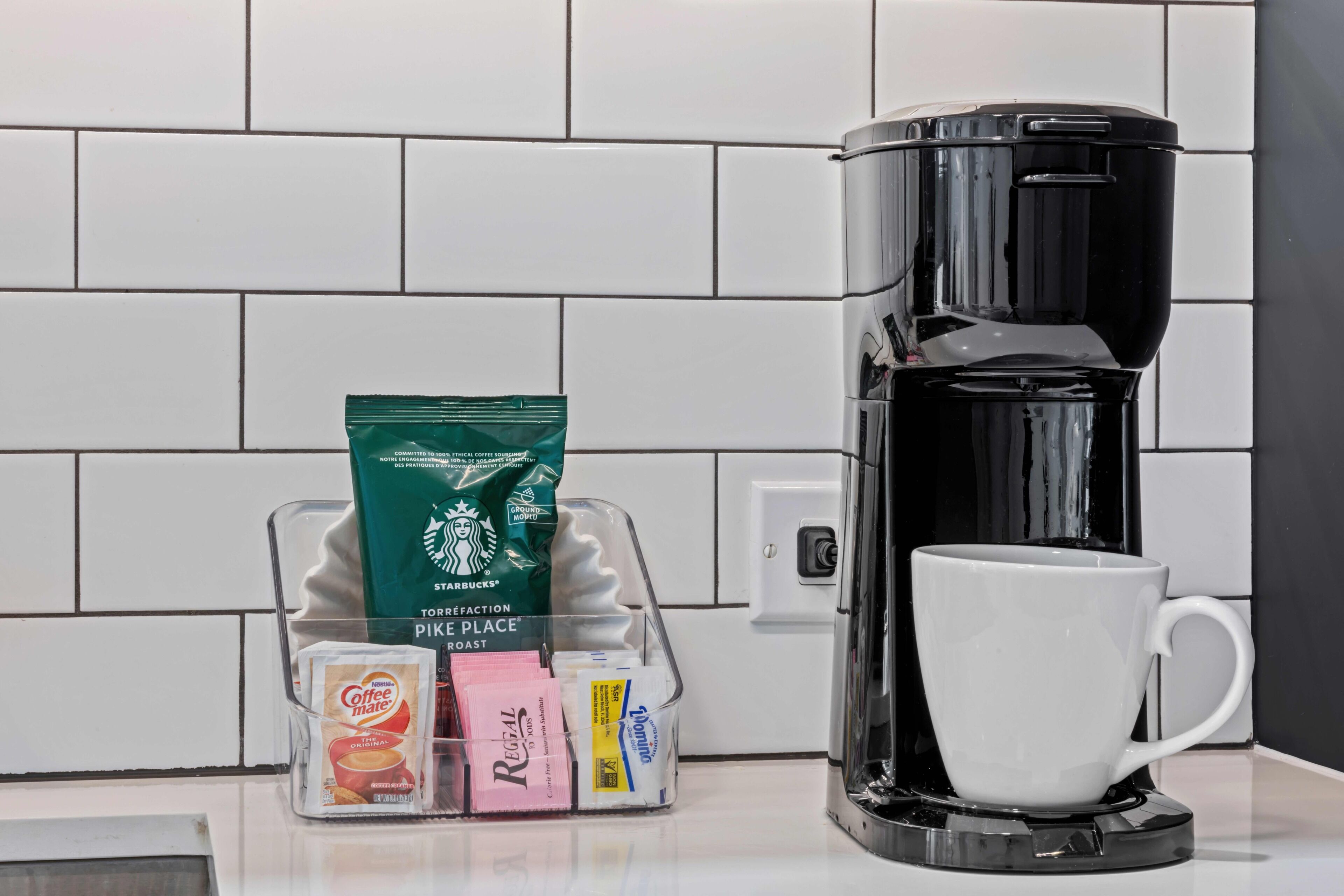 Máquina de café com uma caneca branca ao lado, porta-cápsulas com diversas cápsulas de café, e um saco de café Starbucks.