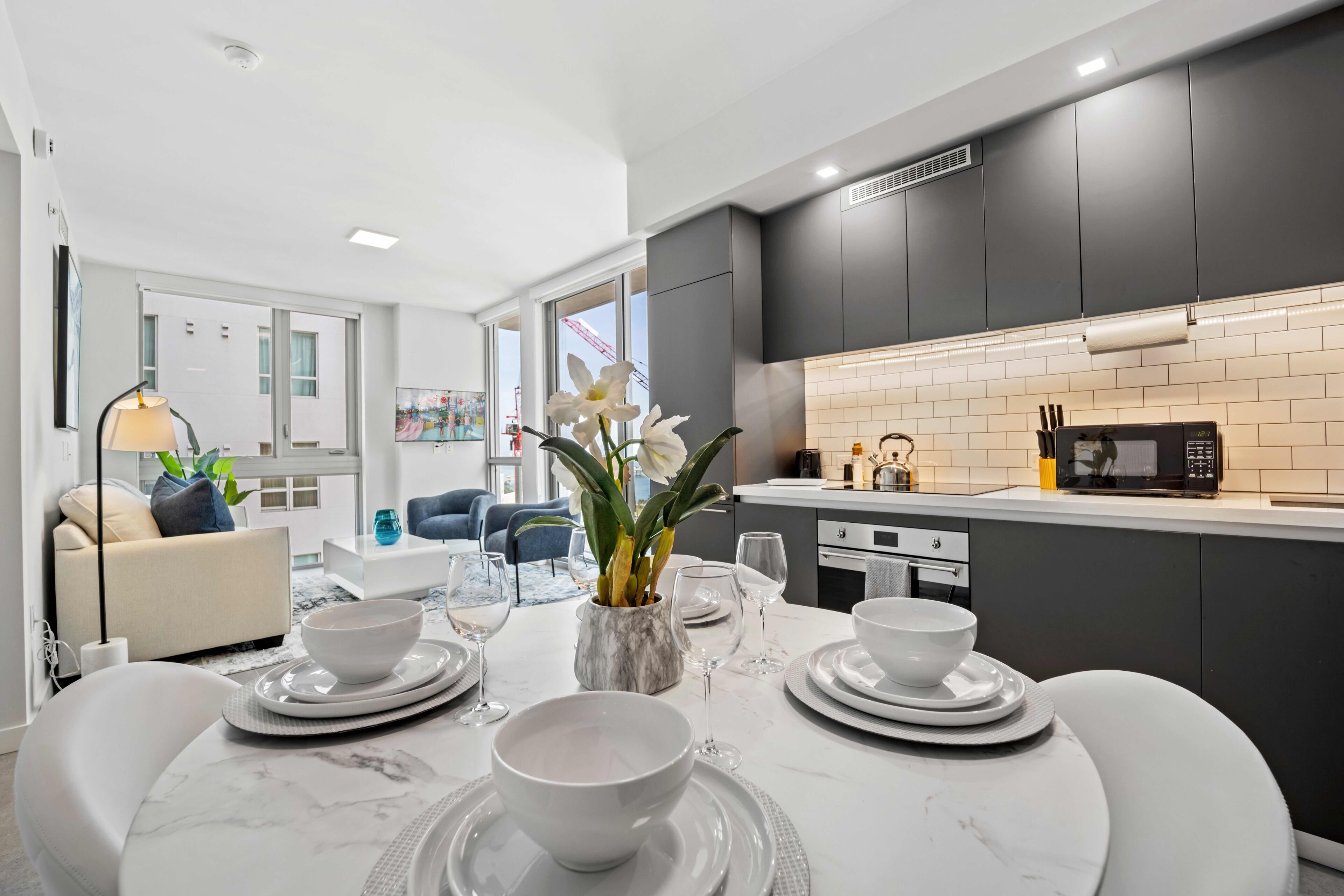 Une cuisine moderne avec des meubles de couleur sombre, un plan de travail blanc et un coin repas agrémenté d’une table en marbre, dressée pour quatre personnes.