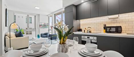 Une cuisine moderne avec des meubles de couleur sombre, un plan de travail blanc et un coin repas agrémenté d’une table en marbre, dressée pour quatre personnes.