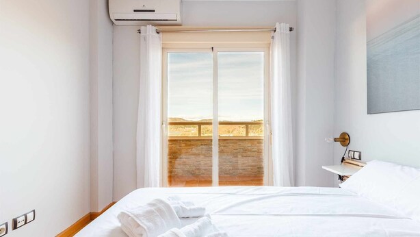 Una camera d'albergo con un letto, asciugamani bianchi, un comodino, una lampada a parete e un'ampia finestra con tende.