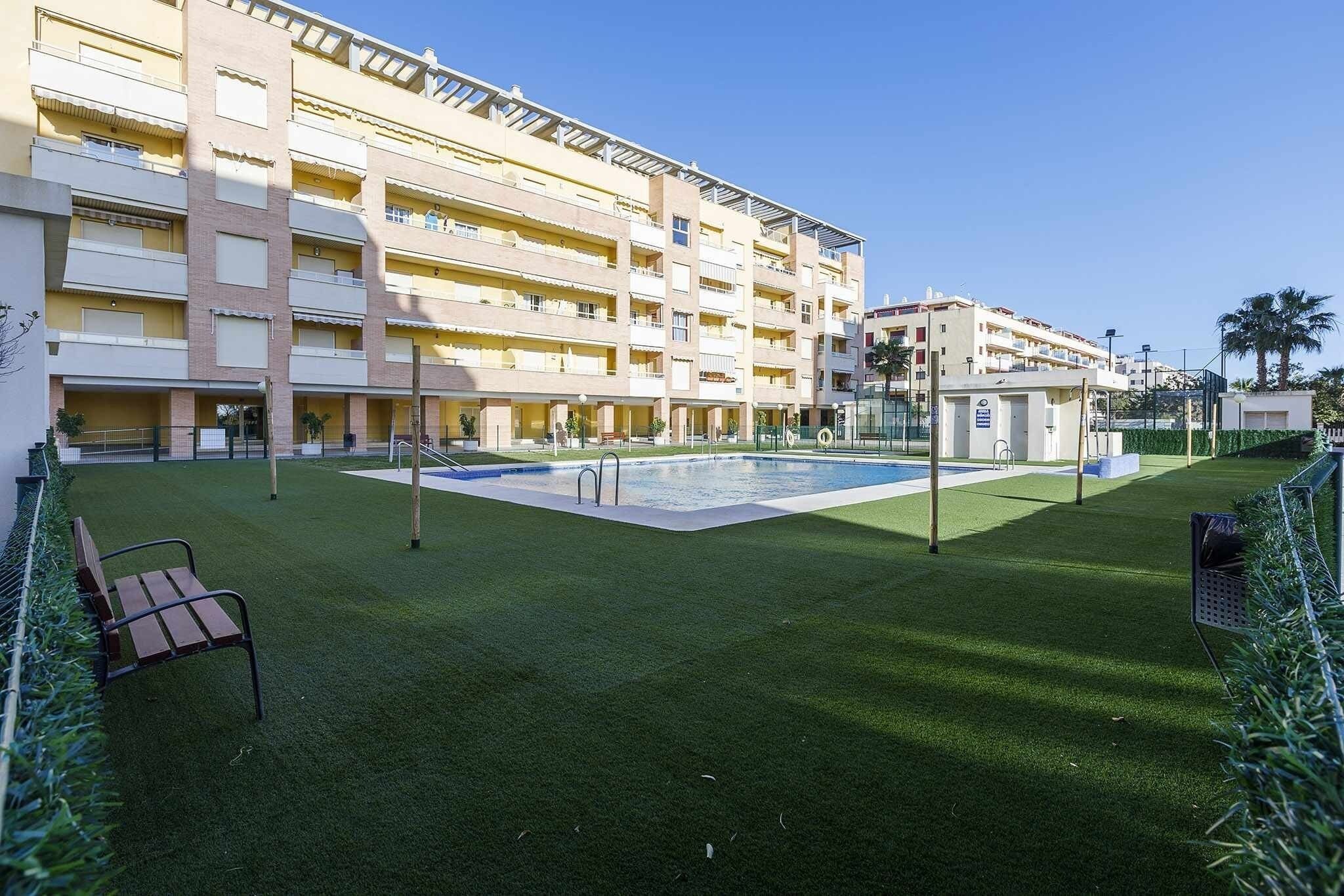 A multi-story hotel with a swimming pool, green lawn, and a bench.