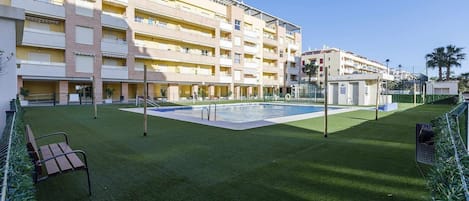 A multi-story hotel with a swimming pool, green lawn, and a bench.
