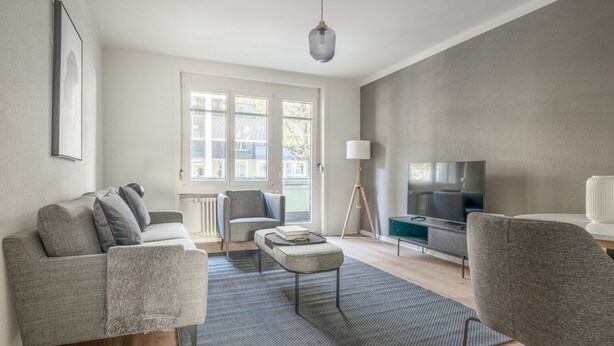 A modern living room with a grey sofa, a small ottoman, a floor lamp, and a TV on a stand. - Mod Wiedikon 2.5 Room w/ Storage & W/D, 1 min to Market, by Blueground (Zurich)