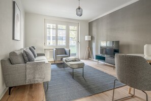 A modern living room with a grey sofa, a small ottoman, a floor lamp, and a TV on a stand. - Mod Wiedikon 2.5 Room w/ Storage & W/D, 1 min to Market, by Blueground (Zurich)